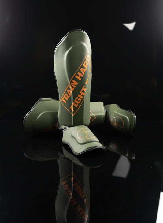 Schienbeinschoner in Military orange aus echtem Leder Ein Schoner  in Vorderansicht der andere quer dahinter liegend, robuster Schutz für Kickboxen